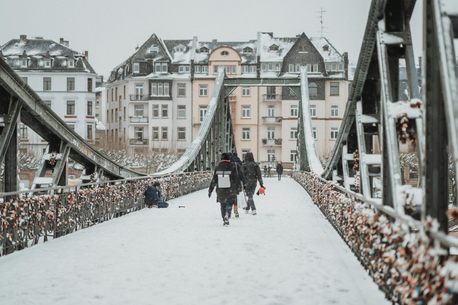 Pedestrians walking across a snow-covered iron footbridge in a historic European city — similar to Copenhagen's Dronning Louise bridge in January