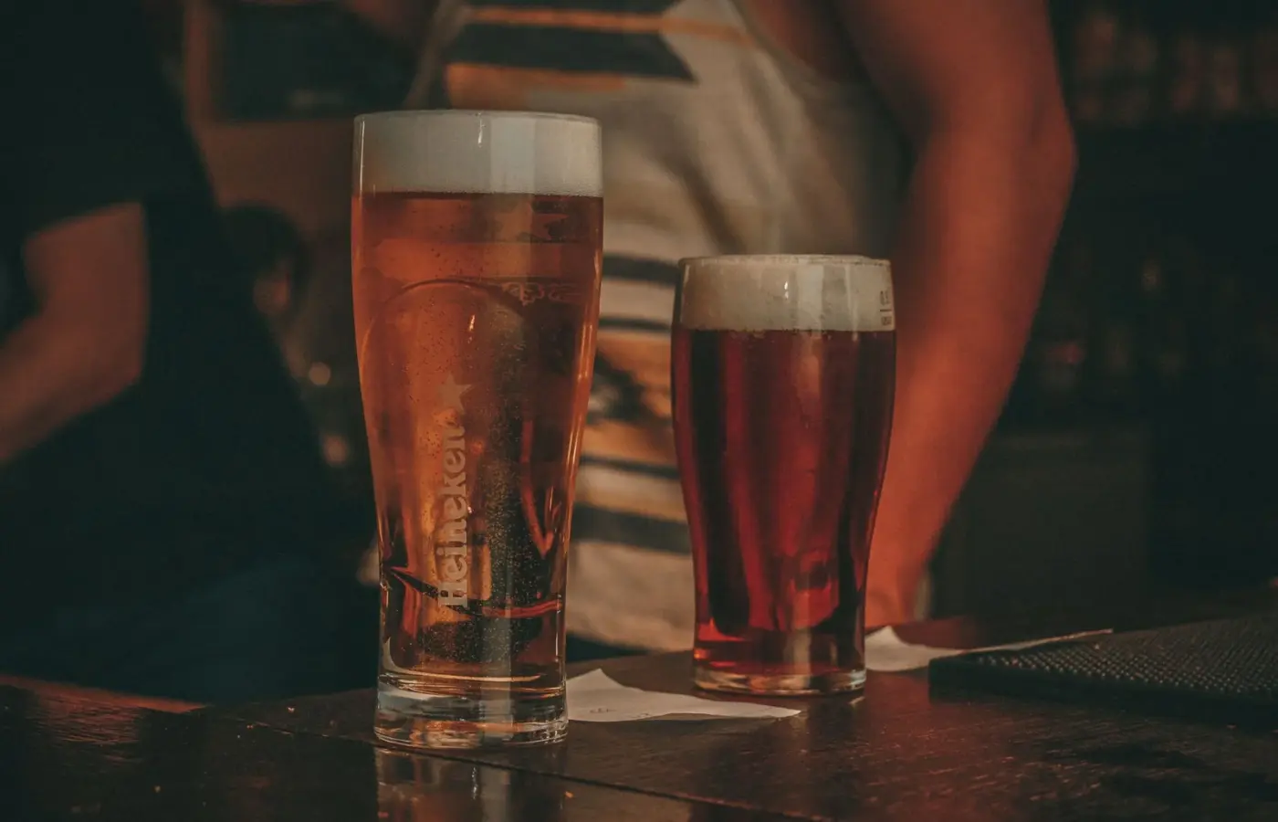 Two glasses of beer standing on a dark wooden bar counter in a dimly lit bar