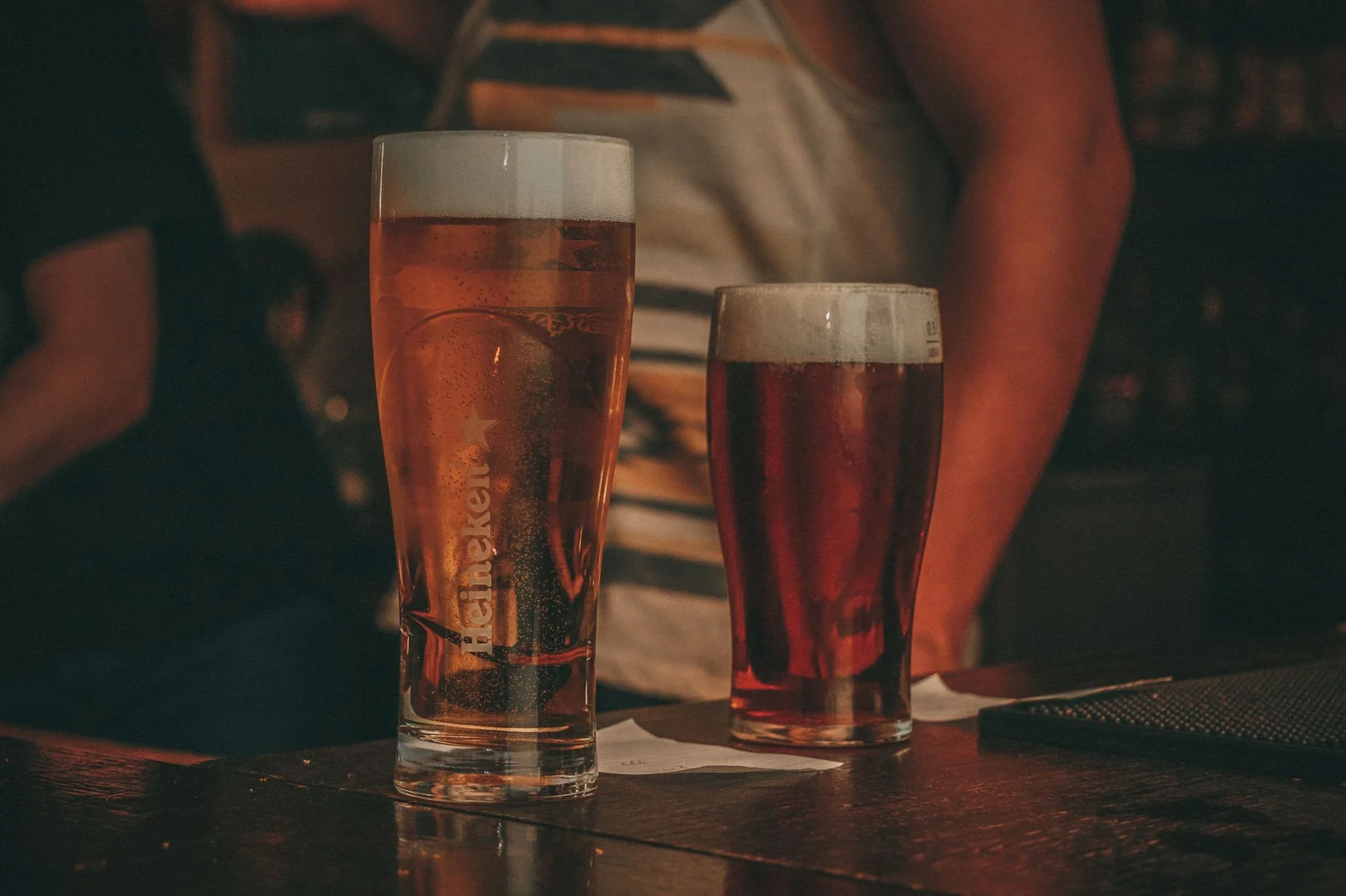 Two glasses of beer standing on a dark wooden bar counter in a dimly lit bar