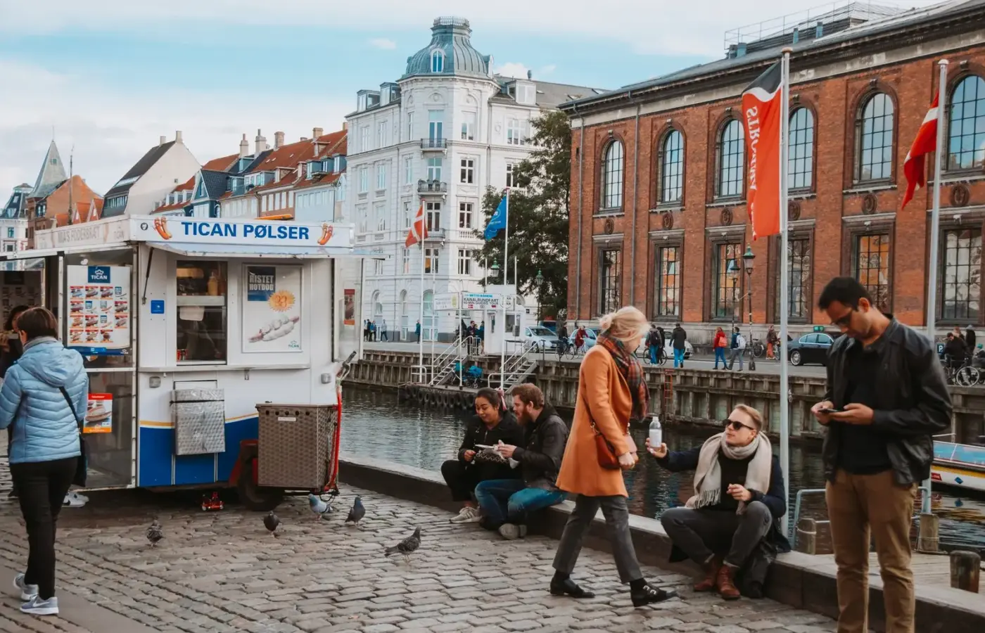 A Danish hot dog van beside a Copenhagen canal with people eating on the quayside