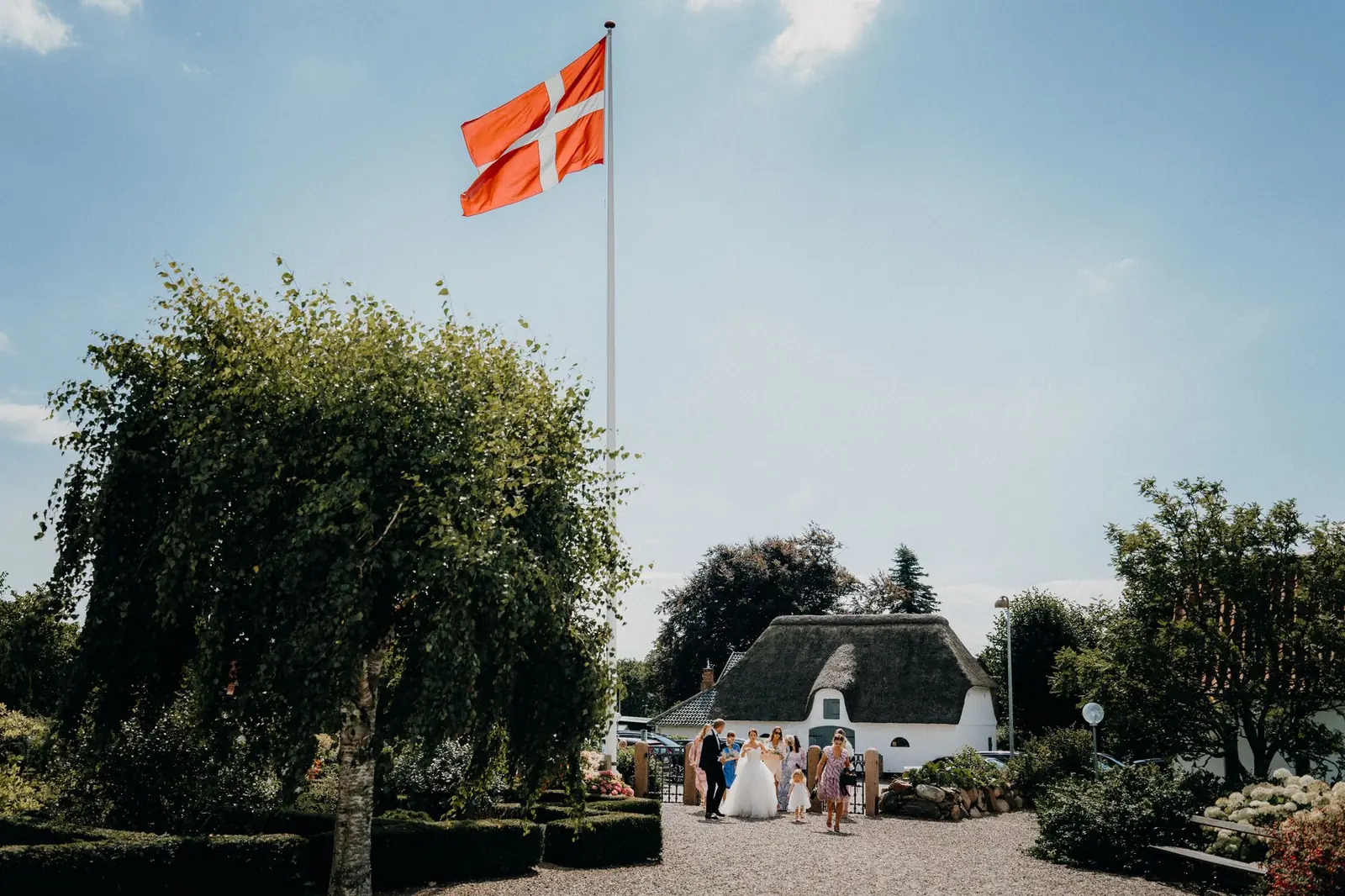 A Danish flag flying above a wedding party outside a thatched farmhouse on a sunny day