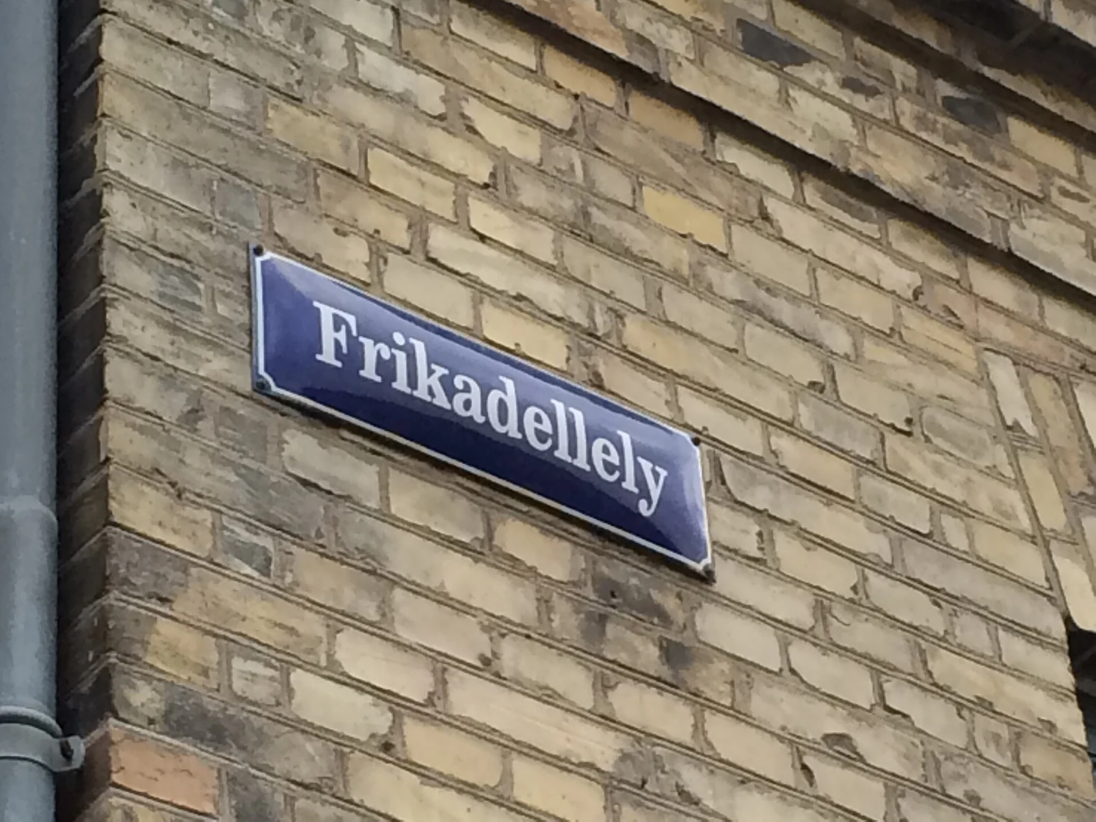 A blue enamel Copenhagen street sign reading Frikadellely on a yellow brick wall
