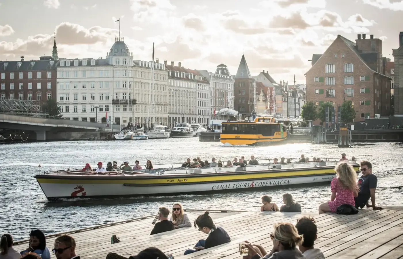 People sitting on a wooden harbour-front deck in the evening sun as a canal tour boat passes in Copenhagen