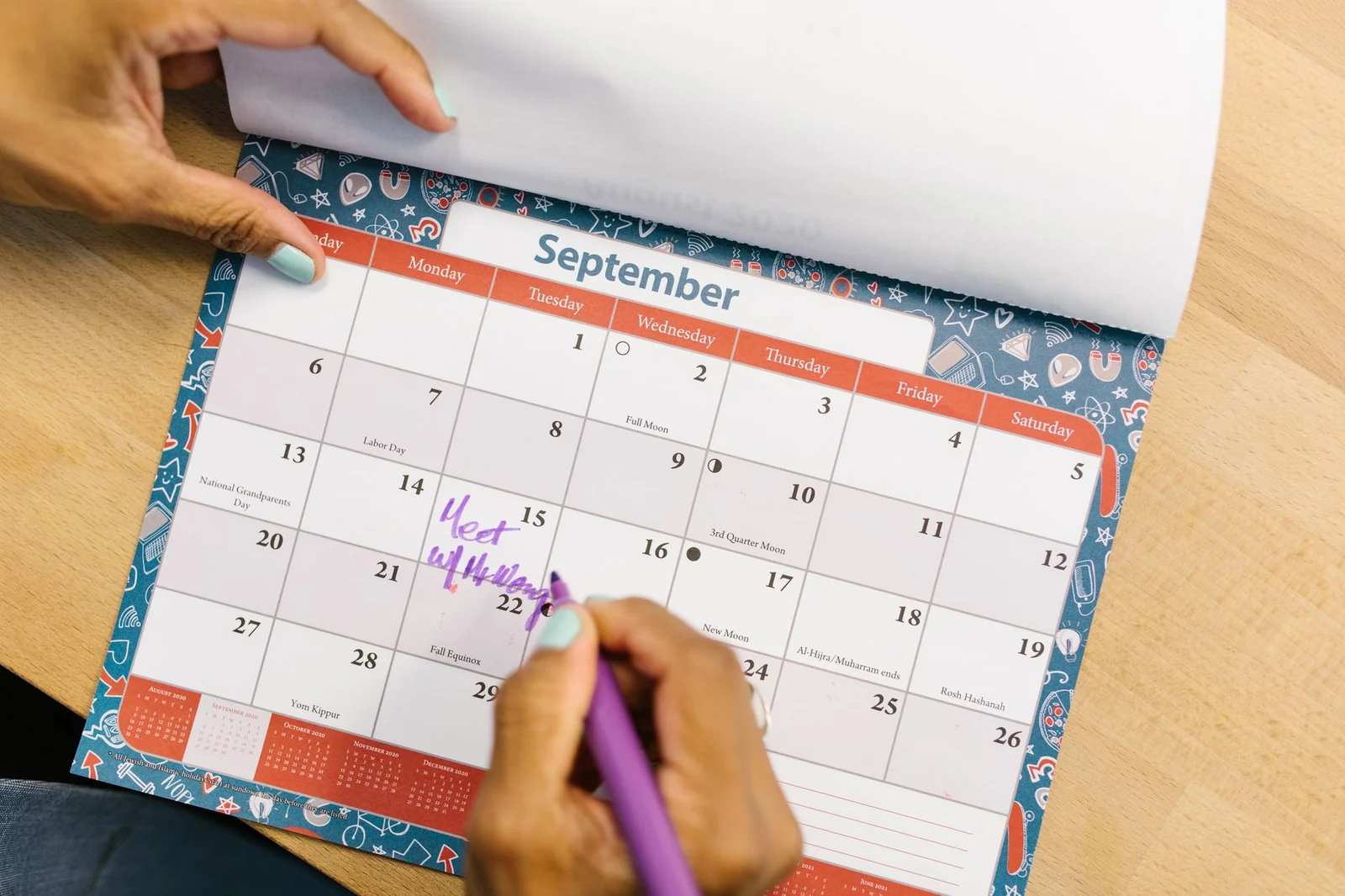 A hand marking dates on a paper wall calendar with a purple pen