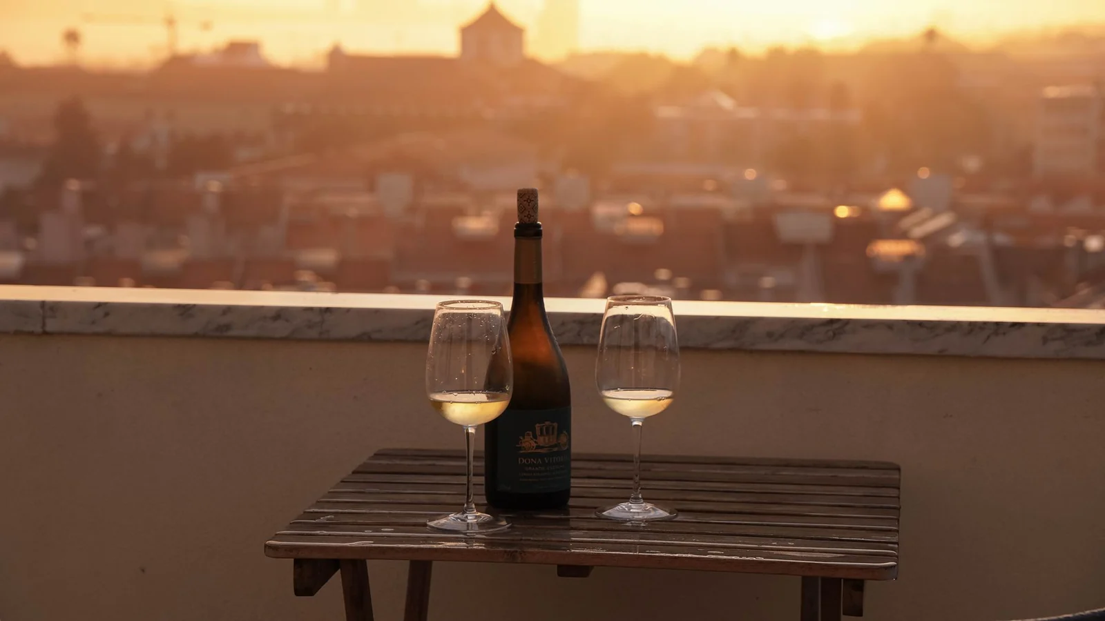 A bottle and two glasses of white wine on a rooftop table above a city at sunset
