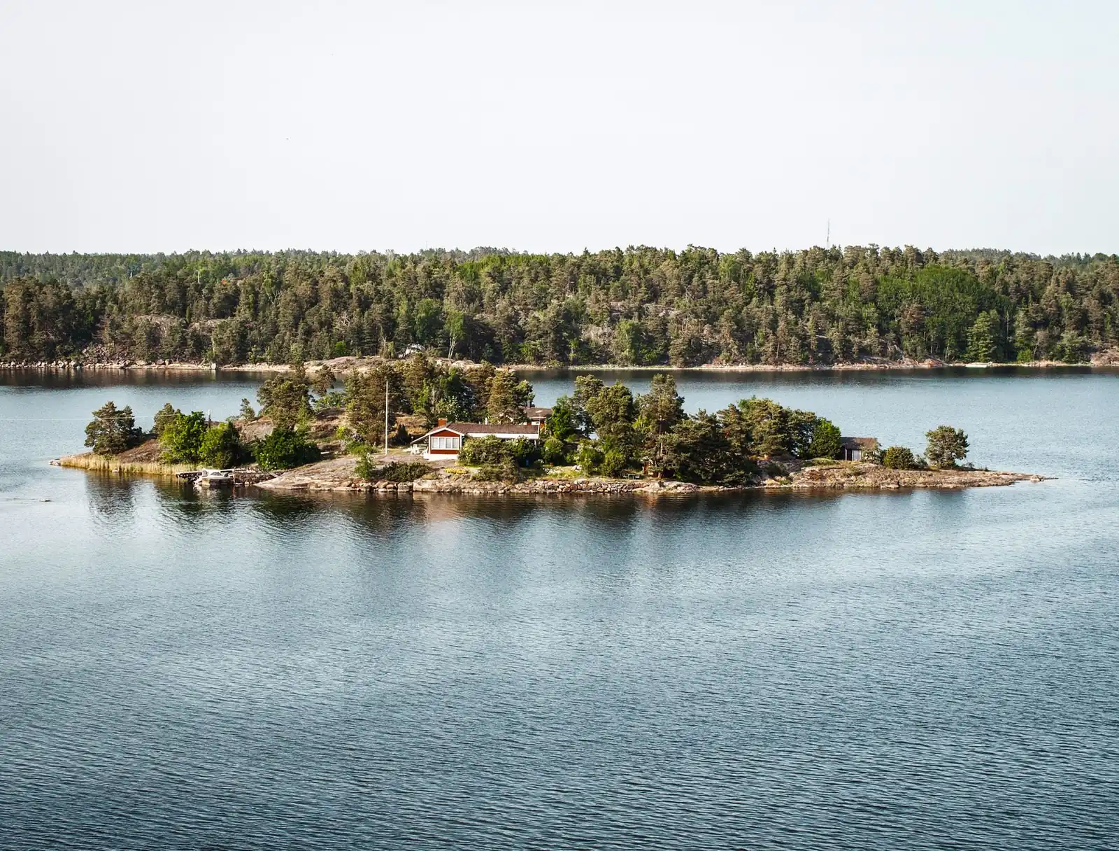 A small wooded island with a red cottage in the calm water of the Stockholm archipelago