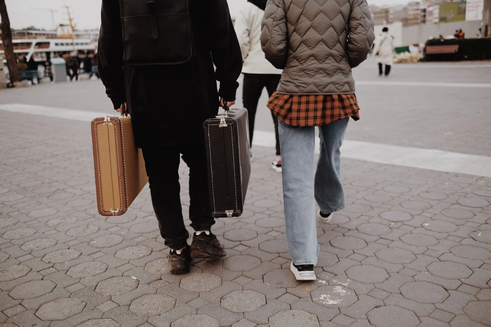 Two travellers seen from behind carrying suitcases across a paved city square
