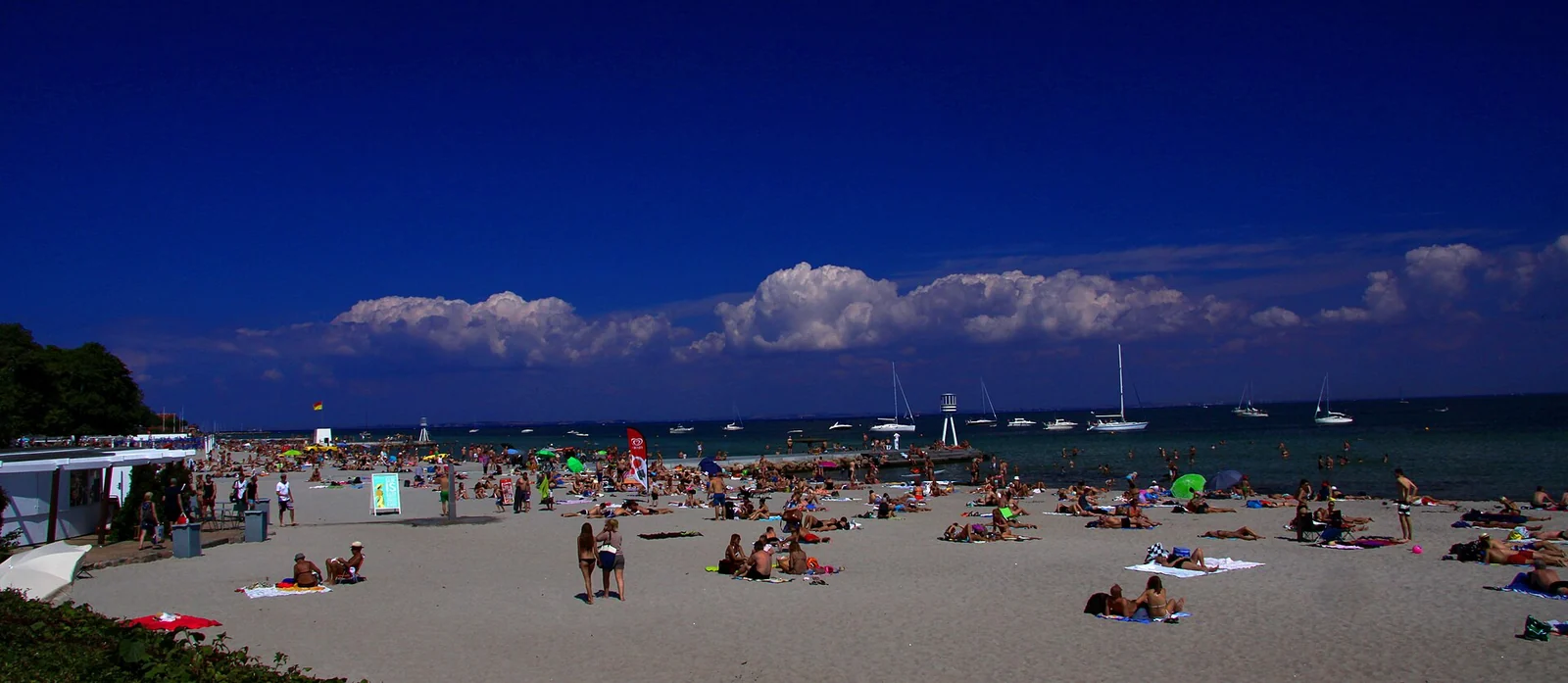 Bellevue beach at Klampenborg packed with sunbathers on a hot summer day, with lifeguard towers and yachts offshore