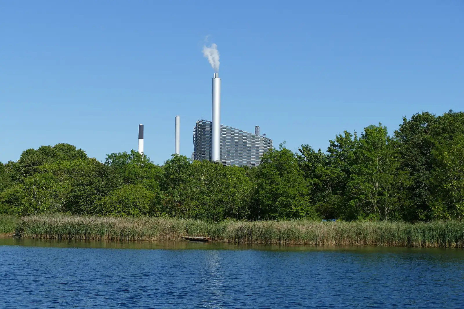 CopenHill, the Amager Bakke waste-to-energy plant with a ski slope on its roof, rising above trees and reeds across the water