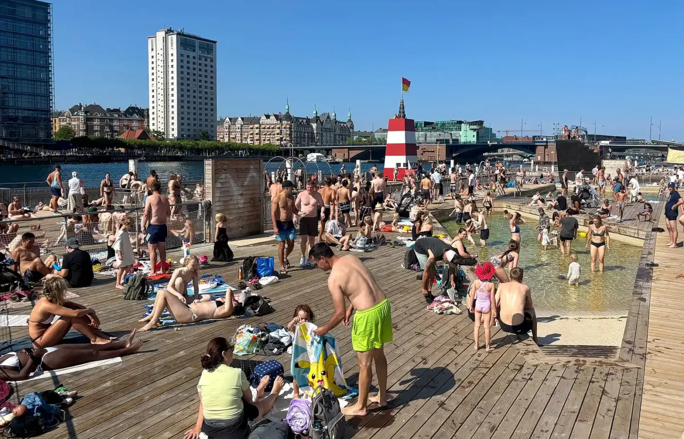 A packed wooden deck and children's pool at Islands Brygge harbour bath on a hot summer day, with the red-and-white lifeguard tower